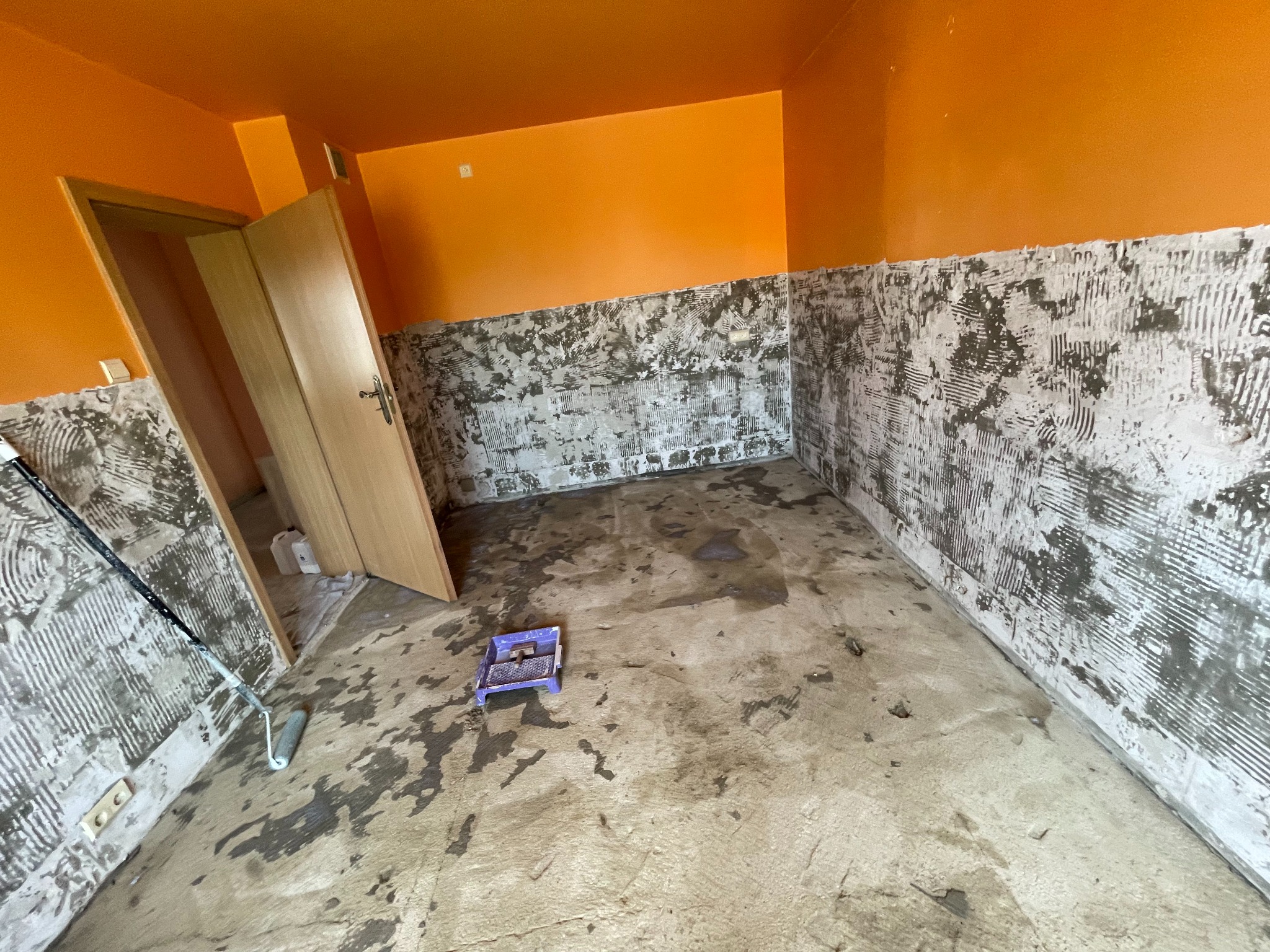 Pomarańczowe wnętrze w trakcie przygotowań do remontu: ściany częściowo pokryte klejem, widoczny wałek malarski i kuweta. Przygotowanie do położenia płytek.