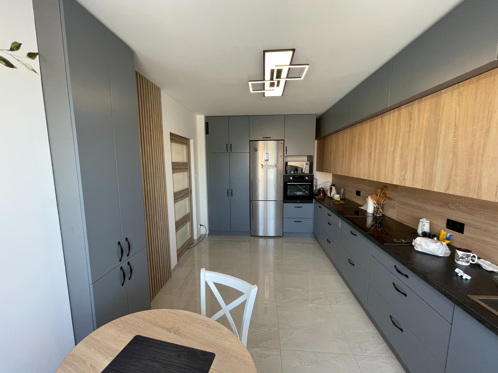 Nowoczesna kuchnia w szarościach i drewnie: zabudowa meblowa, stalowa lodówka, okrągły stół z krzesłem. Jasna podłoga i designerska lampa sufitowa.