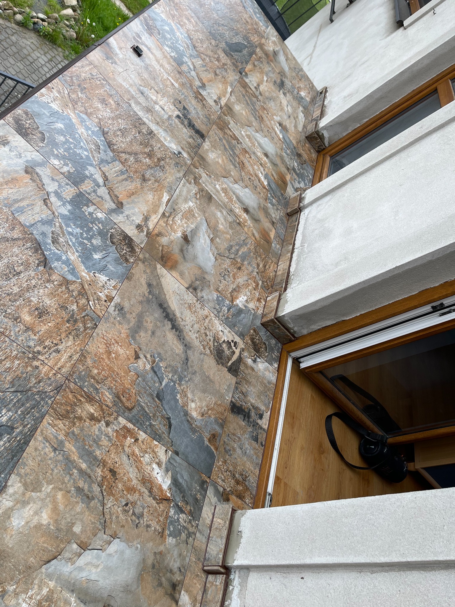Ściana zewnętrzna wyłożona płytkami imitującymi kamień, widok z dołu, z fragmentem okna i balkonu. Płytki w odcieniach szarości, brązu i beżu tworzą naturalny wzór.