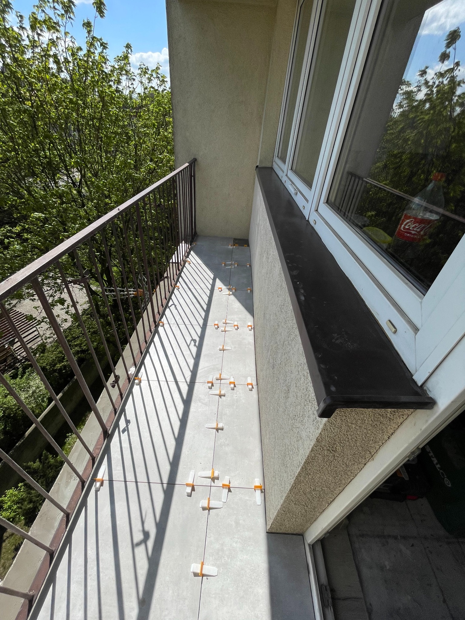 Balkon w trakcie układania płytek podłogowych. Widoczne krzyżyki dystansowe, sznurki wyznaczające linie. Metalowa balustrada i okno z czarnym parapetem.