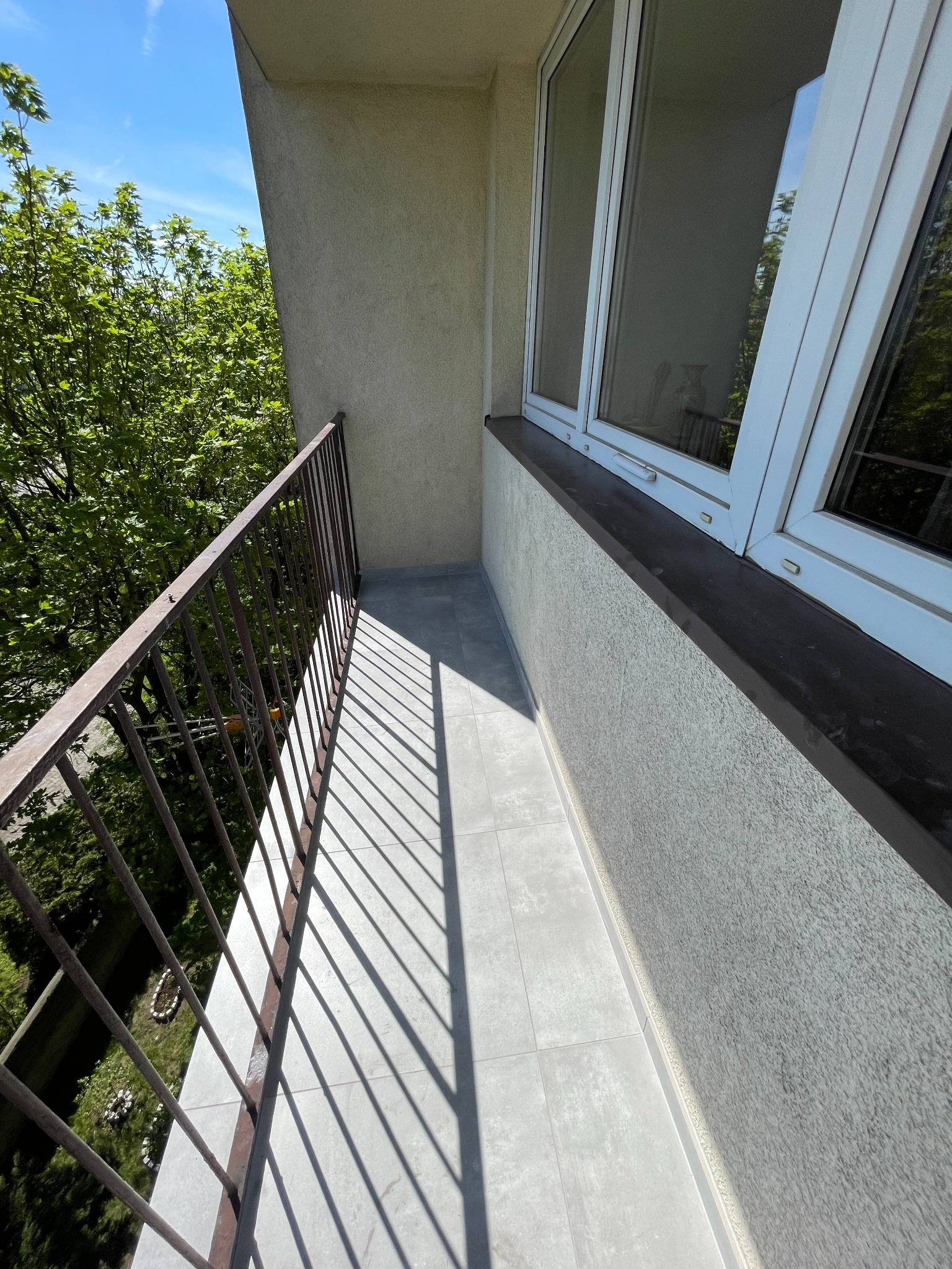 Wykończony balkon z szarą podłogą z płytek, metalową balustradą i widokiem na zieleń. Jasne światło słoneczne podkreśla fakturę ściany i wzór płytek.