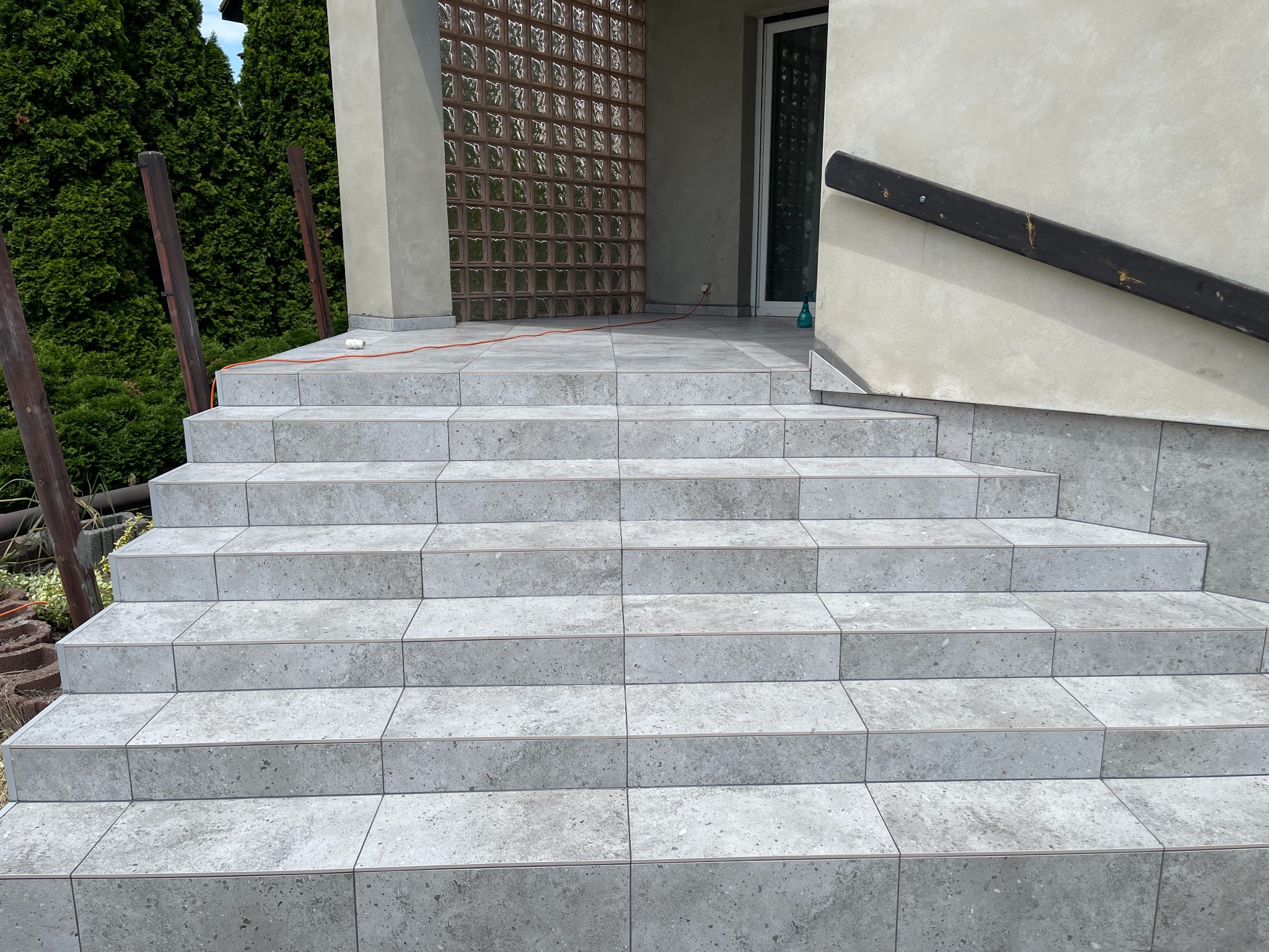 Wykończone płytkami schody wejściowe do domu z szarą fugą, widok z dołu. Na górze widoczny fragment ściany z luksferami i poręczą.