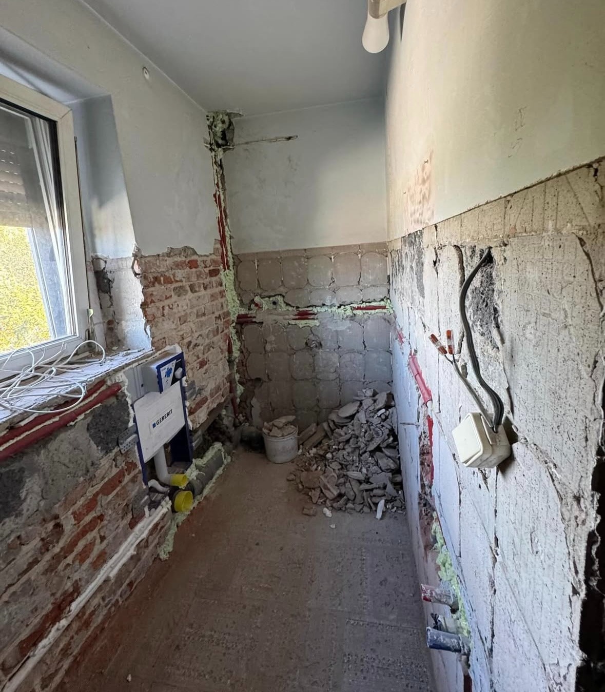 Wnętrze łazienki w trakcie demontażu, widoczne odsłonięte instalacje, fragmenty cegieł i gruzu. Przy oknie biała instalacja sanitarna Geberit.