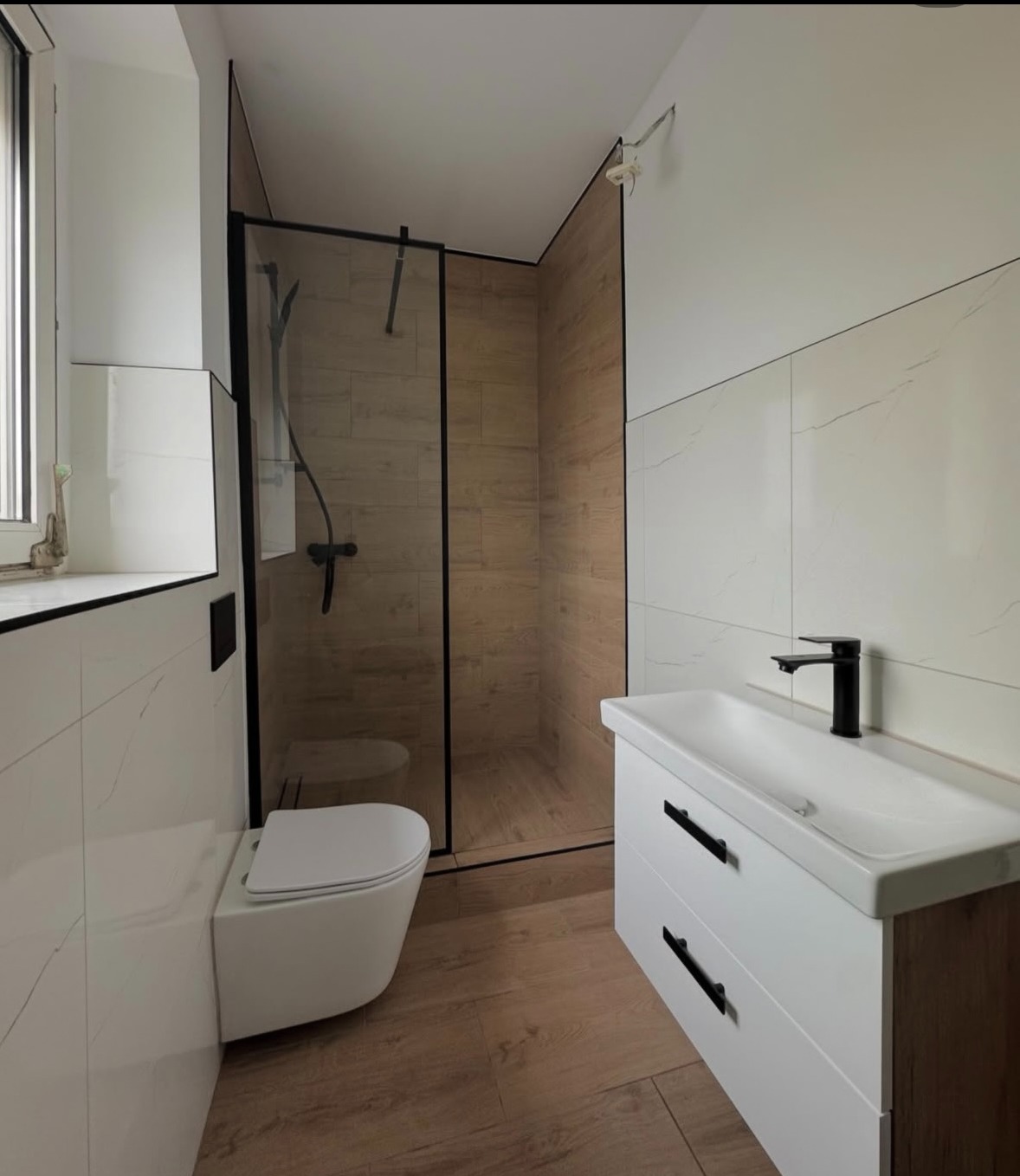 Nowoczesna łazienka z kabiną prysznicową typu walk-in, umywalką z szafką i podwieszaną toaletą. Dominuje biel i drewnopodobne płytki. Czarna armatura.