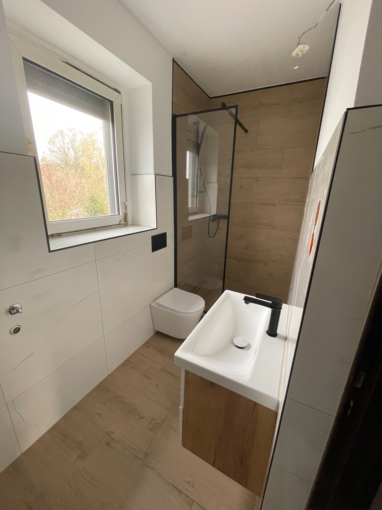 Nowoczesna łazienka po remoncie: białe płytki imitujące marmur, drewnopodobna podłoga, kabina prysznicowa z czarnymi ramami i wisząca umywalka z szafką.