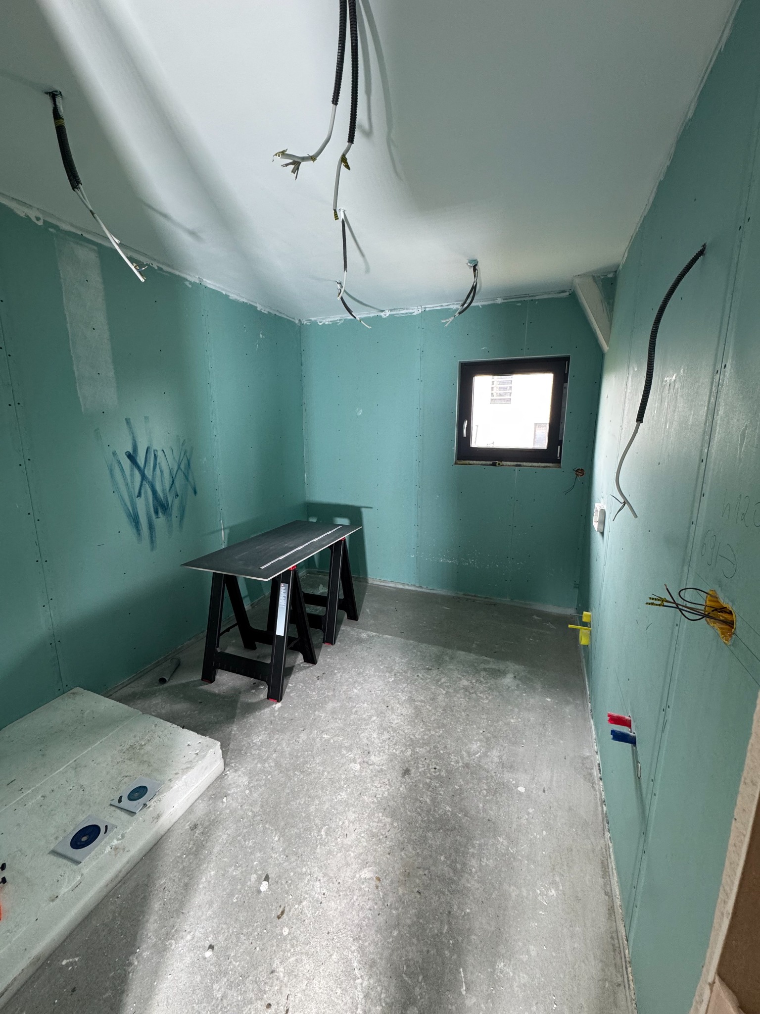 Wnętrze w trakcie prac wykończeniowych: ściany z płyt gipsowo-kartonowych w odcieniach zieleni, widoczne przewody elektryczne, okno i stół roboczy.