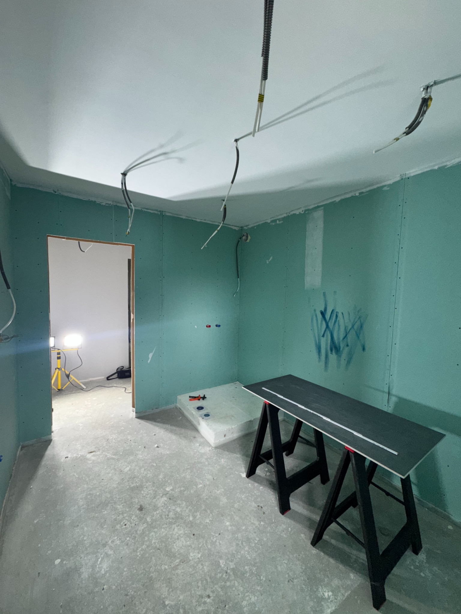 Wnętrze w trakcie prac wykończeniowych: ściany obłożone płytami gipsowo-kartonowymi, widoczne przewody elektryczne, narzędzia i stół roboczy. Oświetlenie halogenowe.