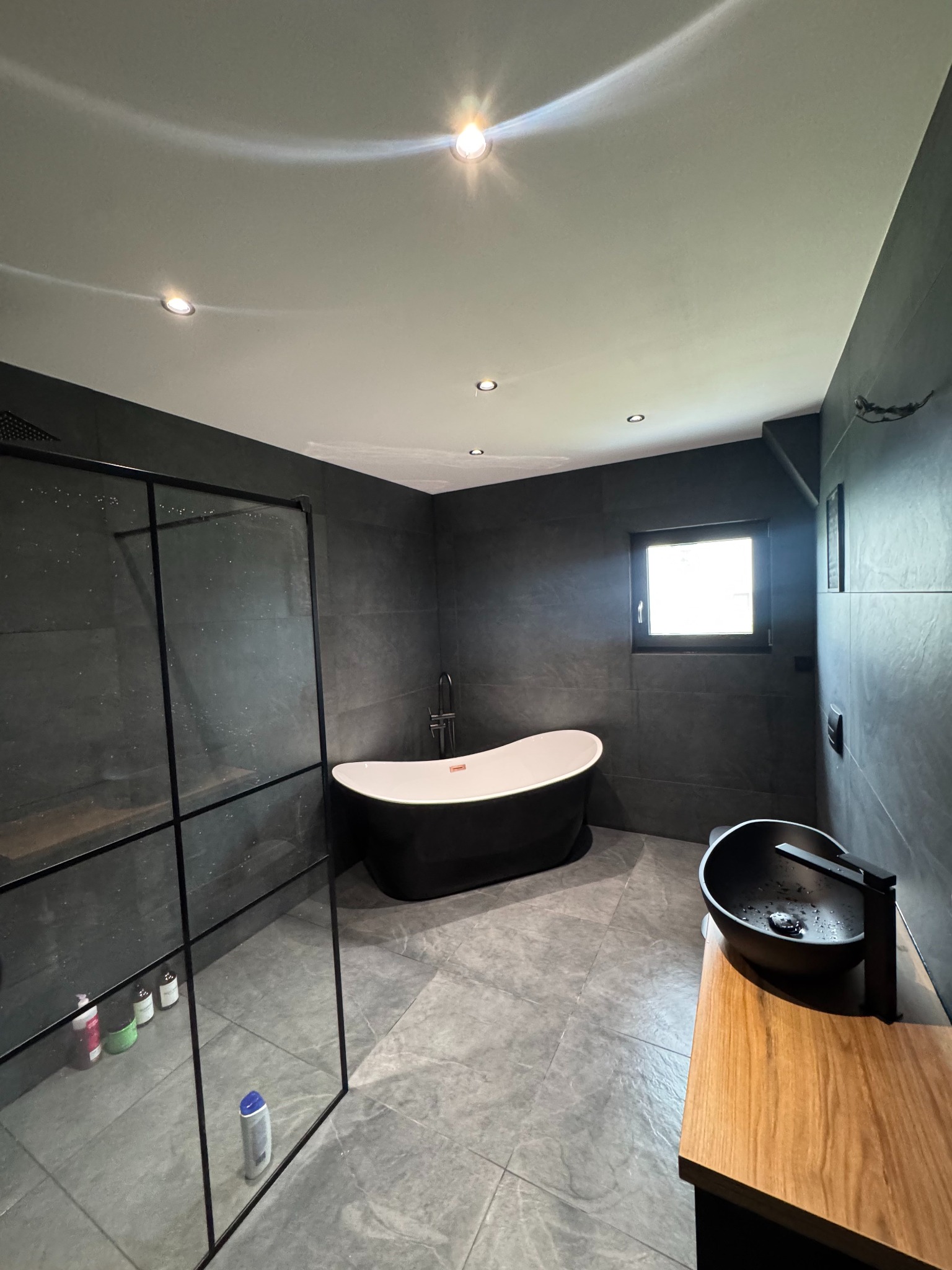 Nowoczesna łazienka z czarną wanną wolnostojącą i kabiną prysznicową typu walk-in. Szare płytki, drewniany blat pod umywalką. Minimalistyczny design, punktowe oświetlenie.
