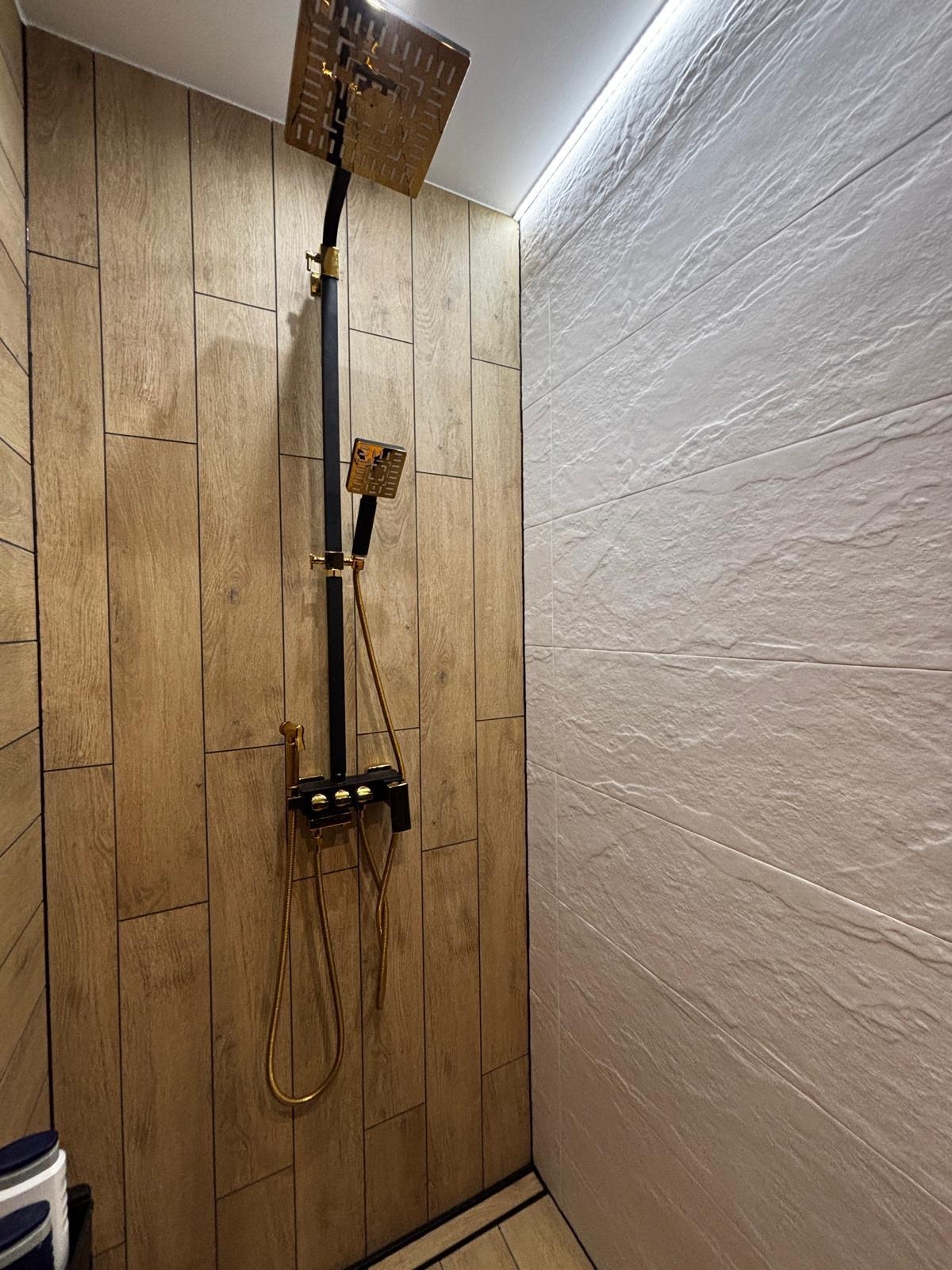 Wykończona kabina prysznicowa z armaturą w kolorze czerni i złota, na ścianach płytki imitujące drewno i kamień. Nowoczesny design, oświetlenie LED.