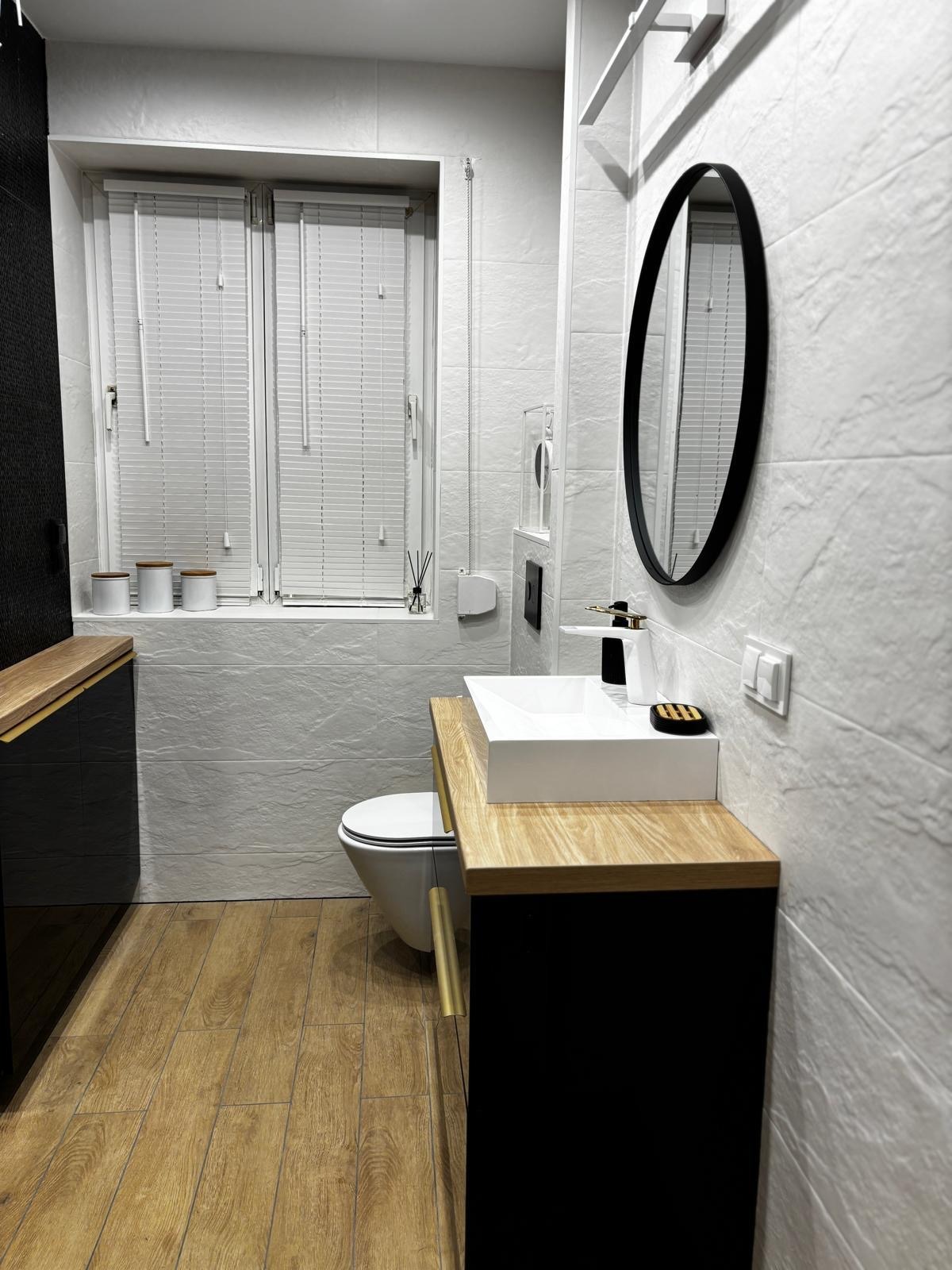 Nowoczesna łazienka po remoncie: drewnopodobna podłoga, białe płytki imitujące kamień, czarne szafki z drewnianym blatem, owalne lustro i okno z żaluzjami.