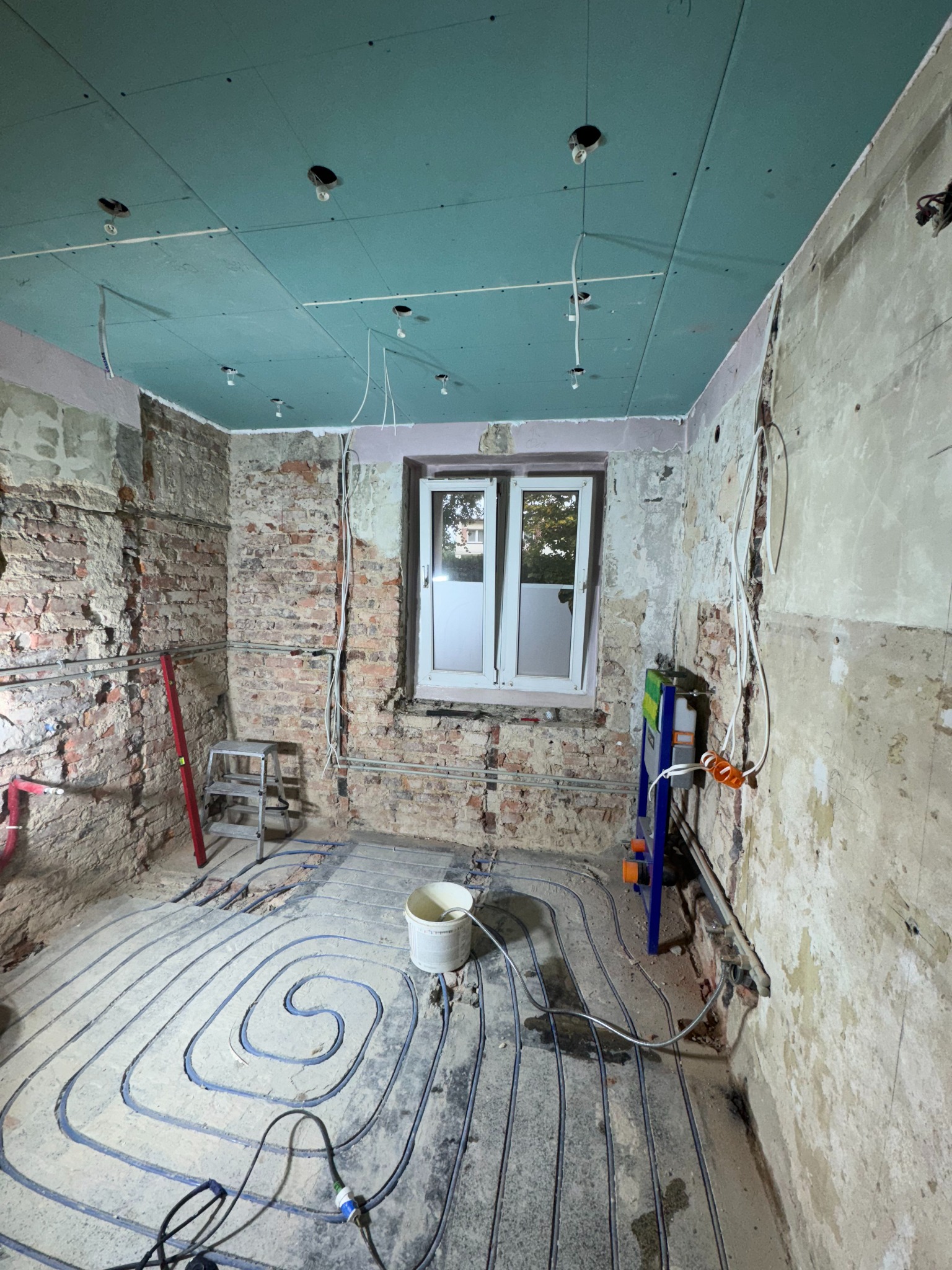 Wnętrze w trakcie remontu: instalacja ogrzewania podłogowego, sufit z płyt karton-gips, odsłonięta cegła, okno. Widoczne narzędzia i elementy instalacji.