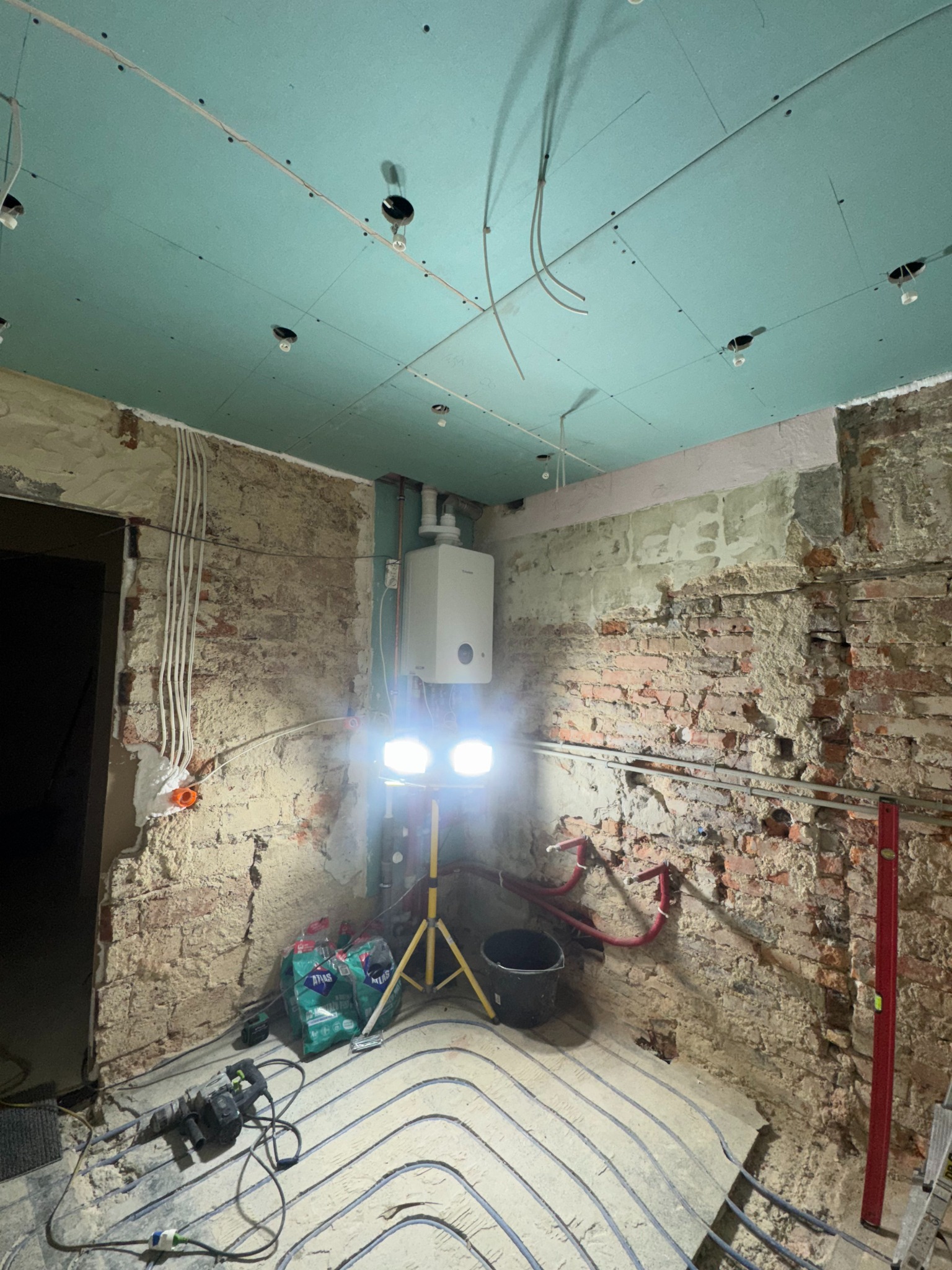 Remont łazienki w trakcie: widoczne instalacje podłogowe, surowa cegła, płyty karton-gips na suficie i bojler. Oświetlenie halogenowe na statywie.