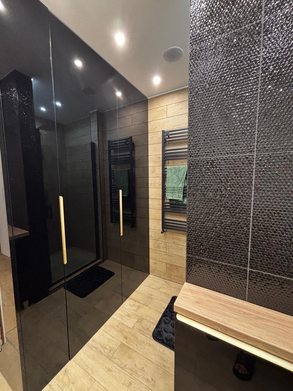 Nowoczesna łazienka po renowacji: czarne, błyszczące płytki na ścianie, drewnopodobne panele, czarny grzejnik drabinkowy z zielonym ręcznikiem i szklana kabina prysznicowa.