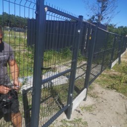 Marta Koceja - Czarny panelowy płot z bramką, montowany przez pracownika z użyciem wkrętarki. W tle drzewa i błękitne niebo.