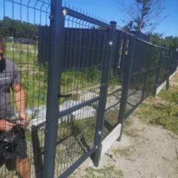 Czarny panelowy płot z bramką, montowany przez pracownika z użyciem wkrętarki. W tle drzewa i błękitne niebo.