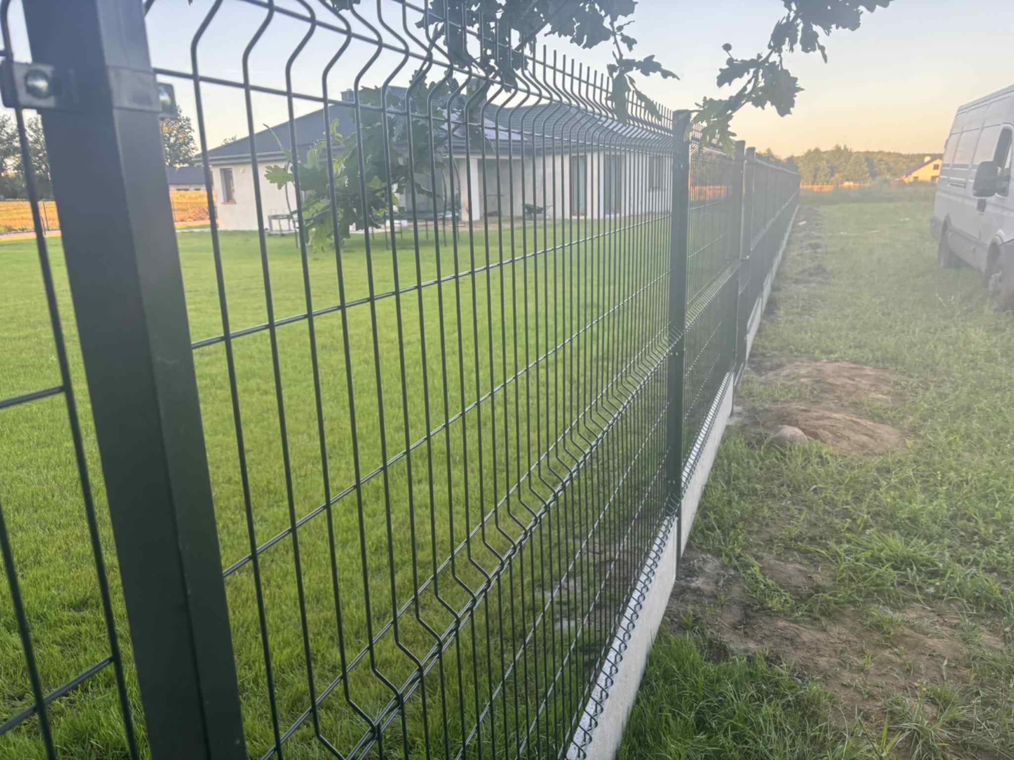 Zamontowane panele ogrodzeniowe na betonowej podmurówce, widok z zewnątrz posesji na tle domu i zieleni. Perspektywa z dołu ukazuje solidność konstrukcji.