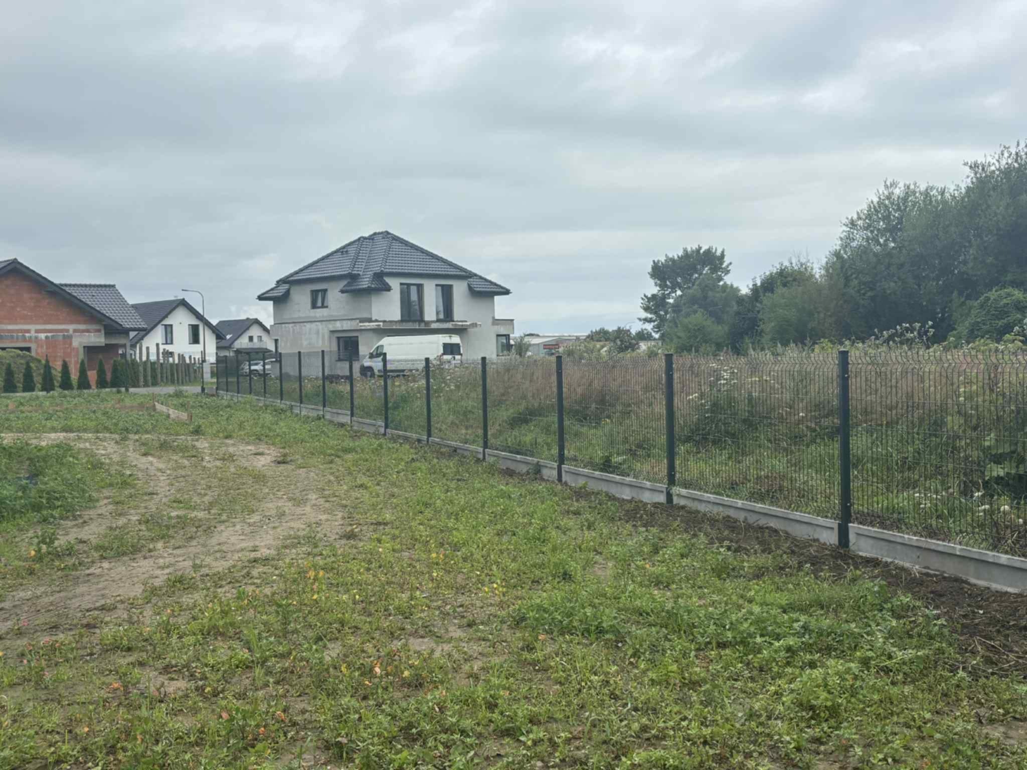 Nowoczesne ogrodzenie panelowe z podmurówką betonową na tle budynków mieszkalnych i zachmurzonego nieba. Widok na posesję z zewnątrz.