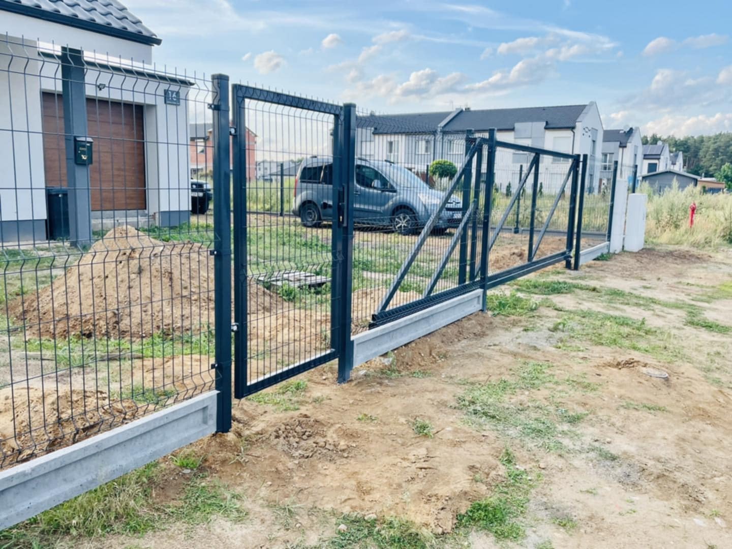 Nowoczesna, przesuwna brama z siatki z metalową ramą, osadzona na betonowej podmurówce. W tle domy jednorodzinne i błękitne niebo z chmurami.