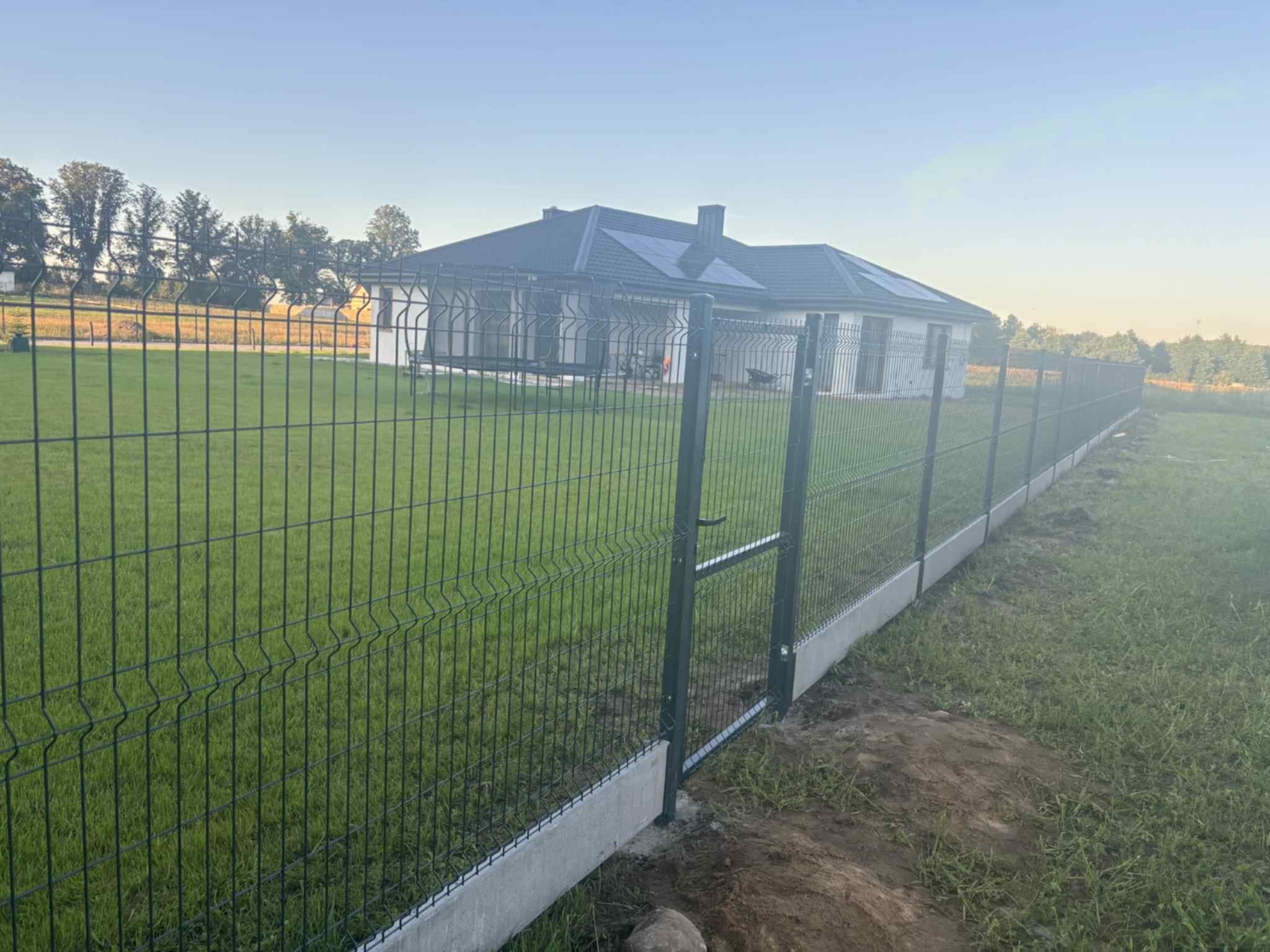 Nowoczesne ogrodzenie panelowe z bramką wejściową, osadzone na betonowej podmurówce, otaczające trawnik przed domem z panelami solarnymi na dachu.