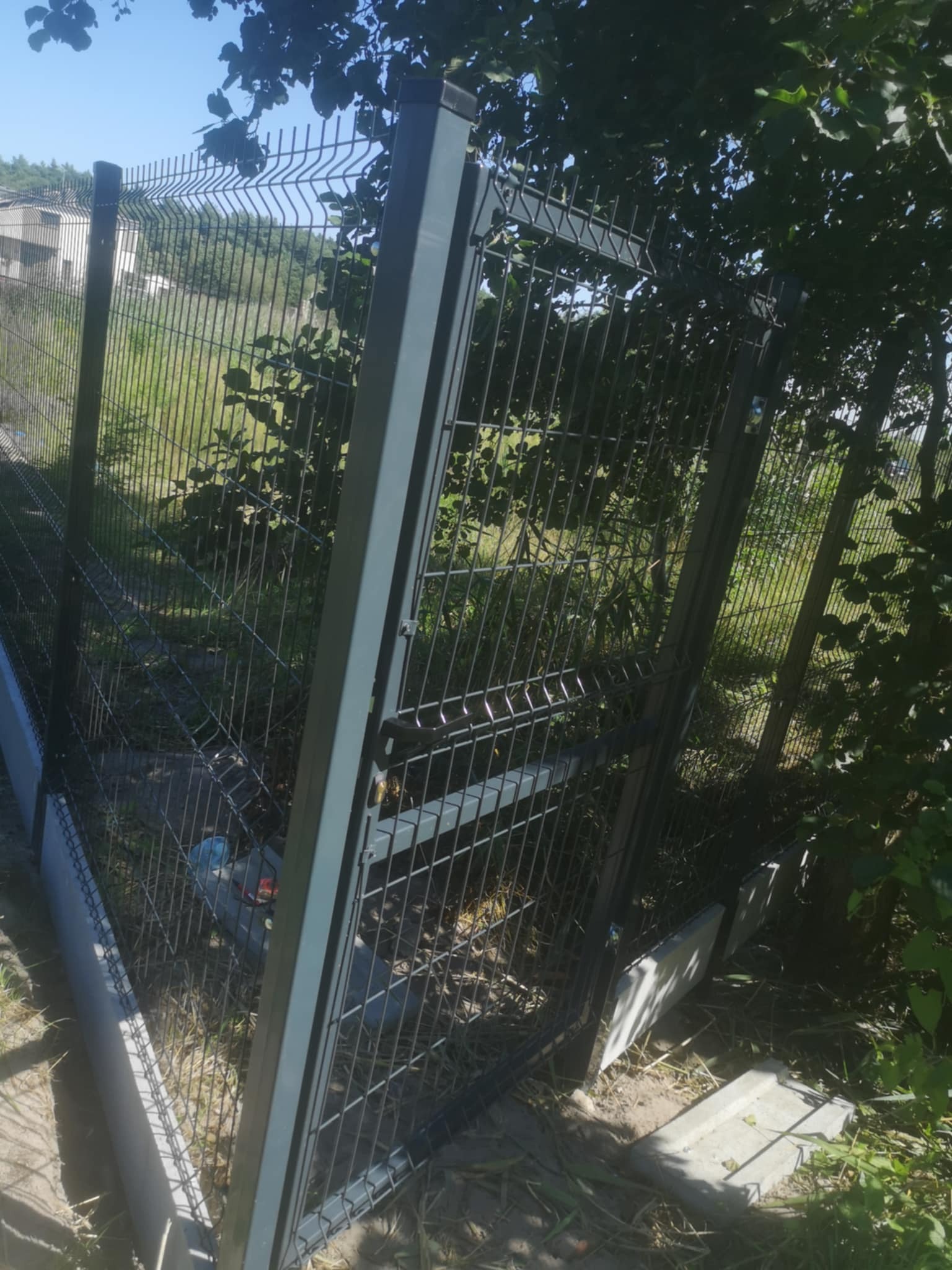 Fragment montażu ogrodzenia panelowego z widocznymi słupkami i siatką, częściowo zasłonięty przez zieleń. Widoczne elementy betonowe i butelka za ogrodzeniem.