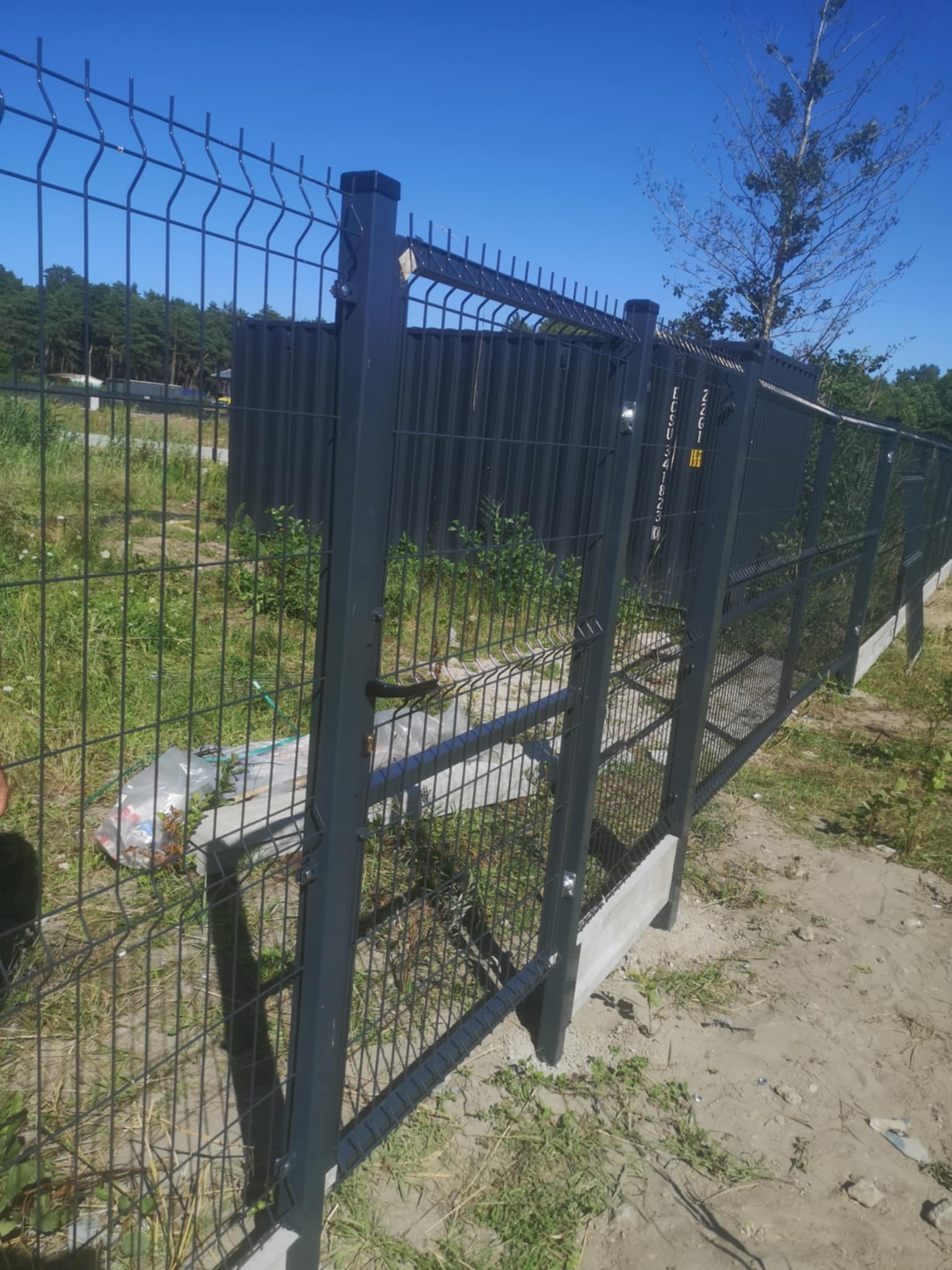 Nowoczesne ogrodzenie panelowe w kolorze grafitowym, z betonową podmurówką, na tle zieleni i błękitnego nieba. Widoczny fragment ziemi i drzewa w oddali.