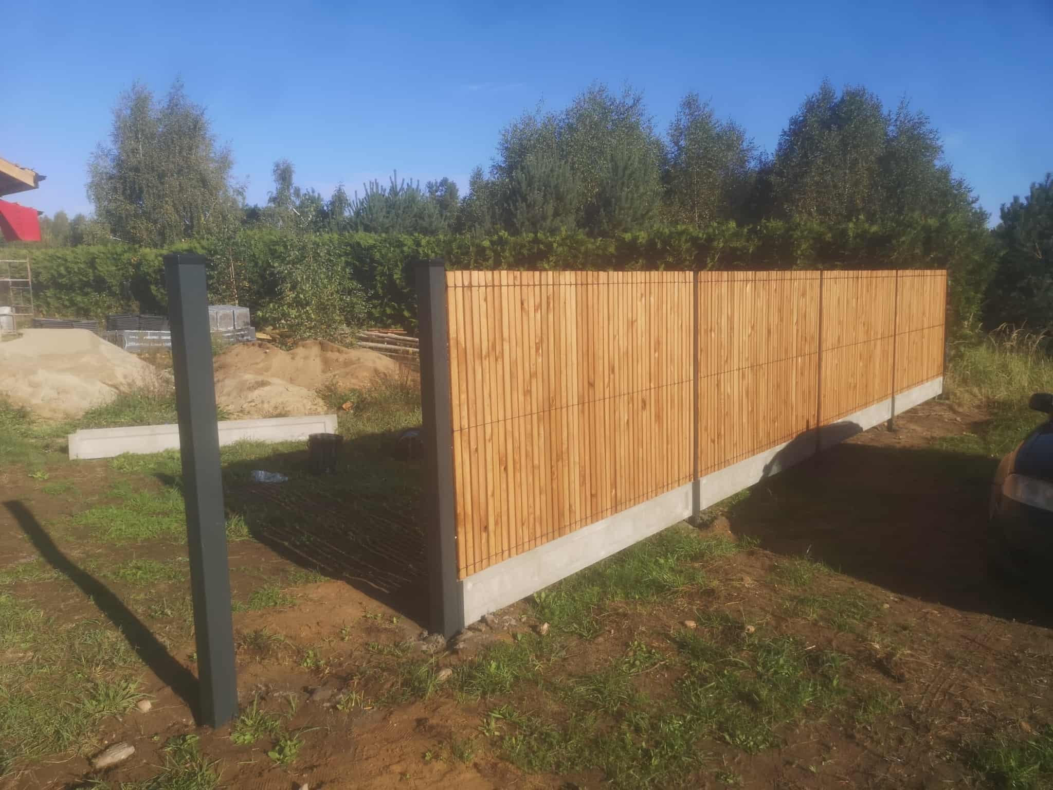 Fragment ogrodzenia z drewnianymi panelami pionowymi i betonową podmurówką, z czarnym słupkiem w tle, na tle zieleni i błękitnego nieba.