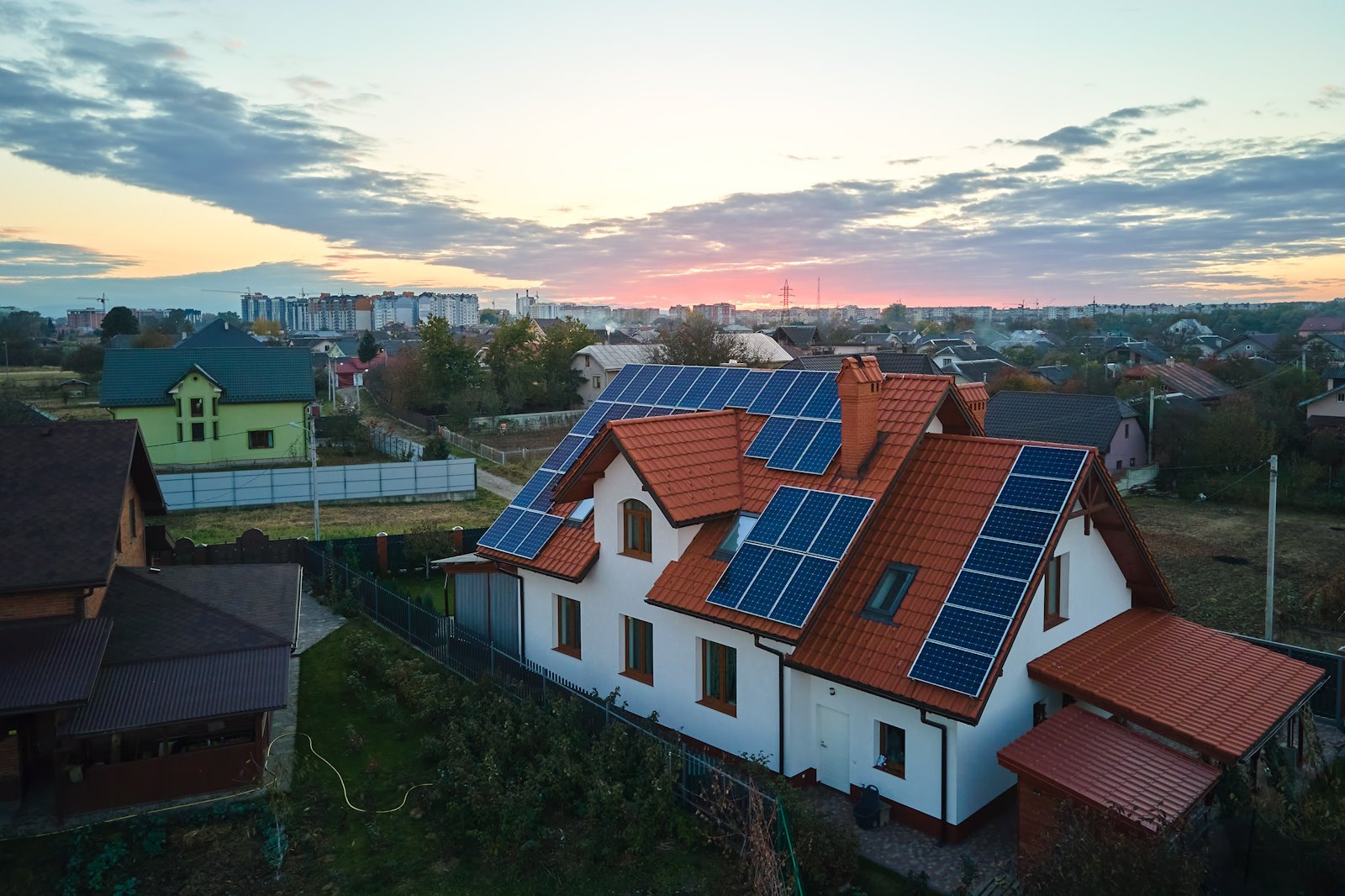 Biały dom z czerwoną dachówką i panelami słonecznymi na dachu, widok z góry o zmierzchu. W tle zabudowa miejska i zachodzące słońce.