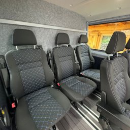 Świerkowe Chatki w Koniakowie - Wnętrze busa z rzędem czarnych foteli z niebieskim wzorem rombów, wykończonych białą nicią. Widoczne szare obicie ścian i drewnopodobna podłoga z aluminiowymi szynami.