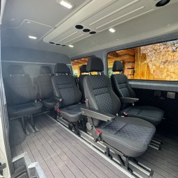 Świerkowe Chatki w Koniakowie - Wnętrze busa z zamontowanymi, regulowanymi fotelami z czarną tapicerką w romby z niebieskim szwem. Podłoga wyłożona panelami imitującymi drewno. Widok z perspektywy wnętrza.