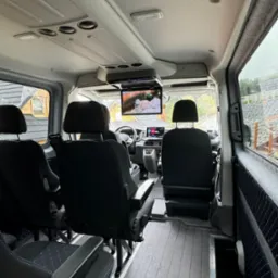 Wnętrze luksusowego busa z czarnymi skórzanymi siedzeniami, drewnianą podłogą i ekranem TV zamontowanym pod sufitem, widok z tyłu pojazdu.