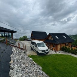 Świerkowe Chatki w Koniakowie - Jasny bus na tle drewnianych domków z czarnymi dachami, otoczonych zielenią i kamieniami. Pochmurne niebo. Widok z dołu, budzący zaufanie.