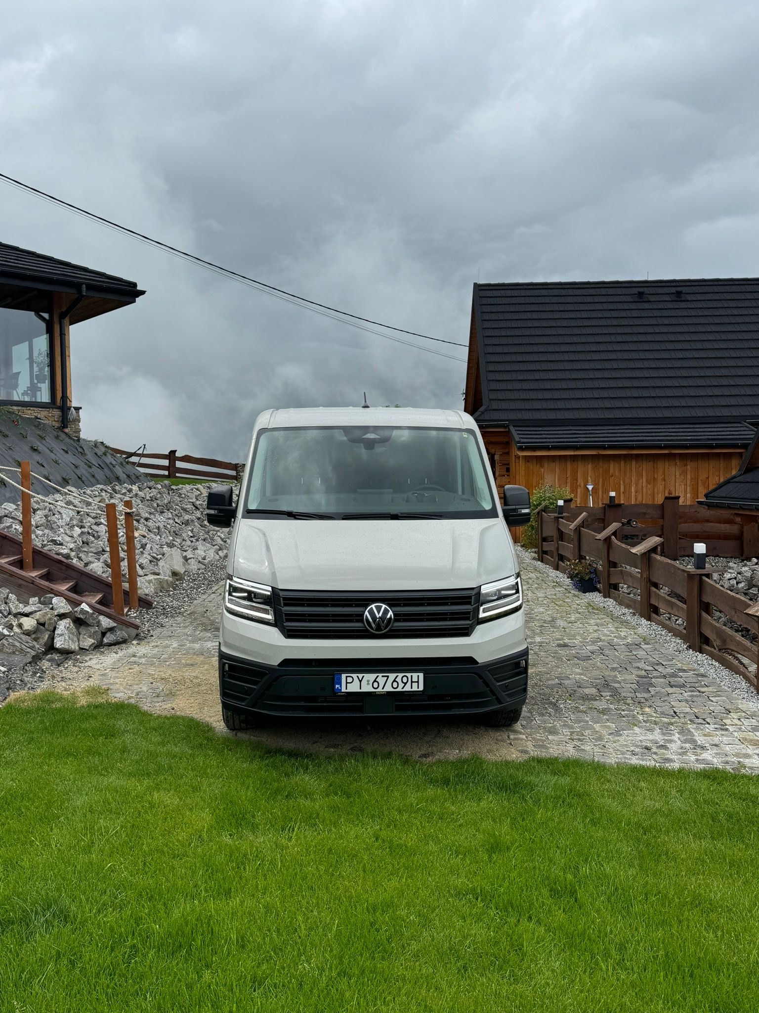 Biały bus Volkswagen na brukowanym podjeździe, górzysty krajobraz w tle, pochmurne niebo. Tablica rejestracyjna PY 6769H.