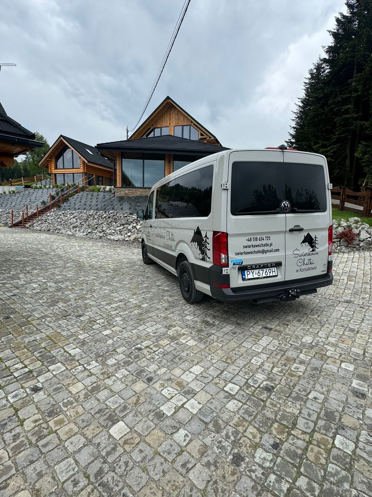 Jasny bus VW Crafter z logotypem 'Świerkowe Chatki' na tle drewnianych domków i brukowanej nawierzchni. Tablica rejestracyjna: PY 6769H.