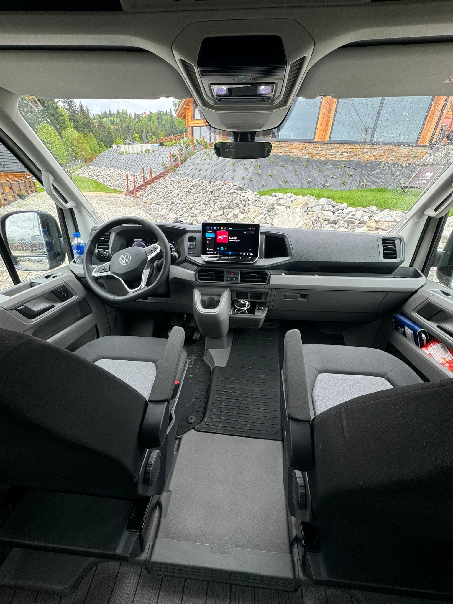 Wnętrze busa VW z widokiem na deskę rozdzielczą i fotele, za szybą dom w górach. Nowoczesny kokpit z ekranem dotykowym i widok na malowniczy krajobraz.
