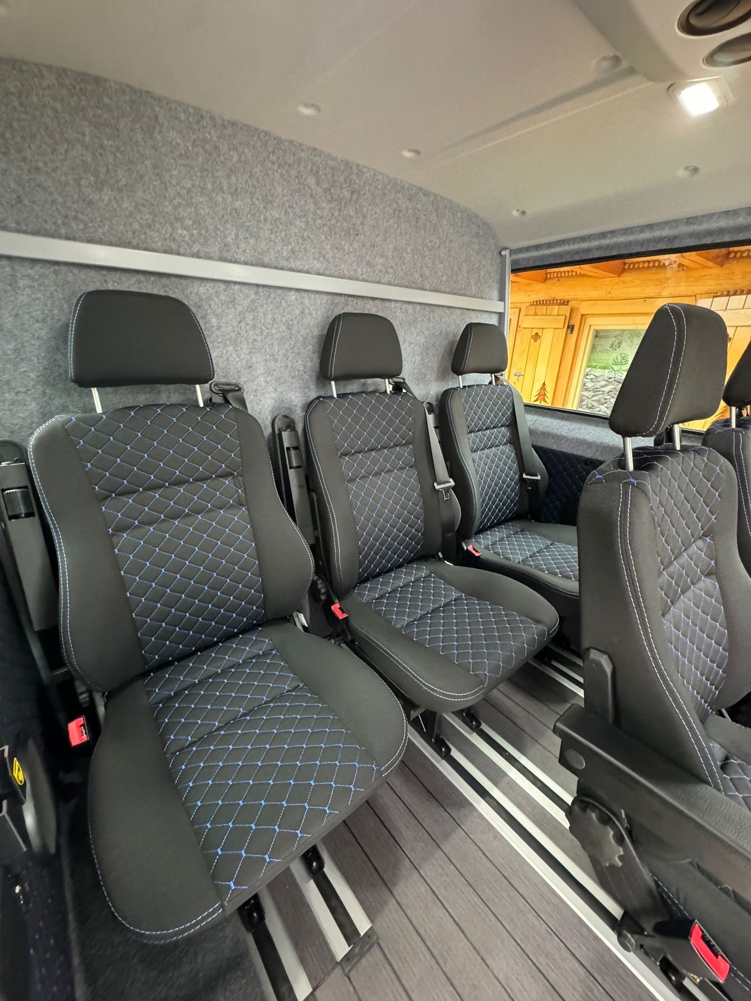 Wnętrze busa z rzędem czarnych foteli z niebieskim wzorem rombów, wykończonych białą nicią. Widoczne szare obicie ścian i drewnopodobna podłoga z aluminiowymi szynami.