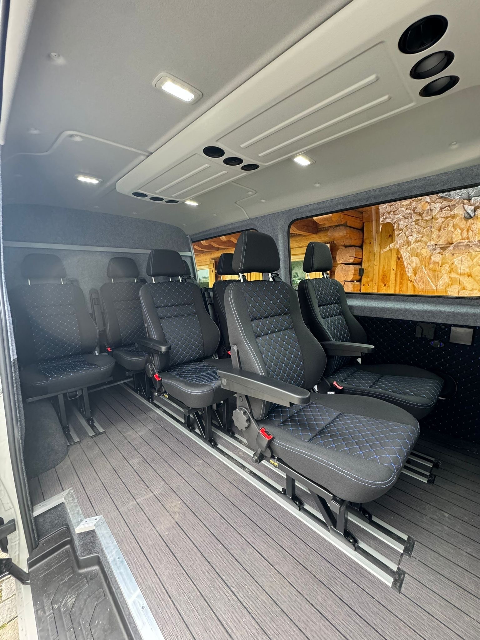 Wnętrze busa z zamontowanymi, regulowanymi fotelami z czarną tapicerką w romby z niebieskim szwem. Podłoga wyłożona panelami imitującymi drewno. Widok z perspektywy wnętrza.