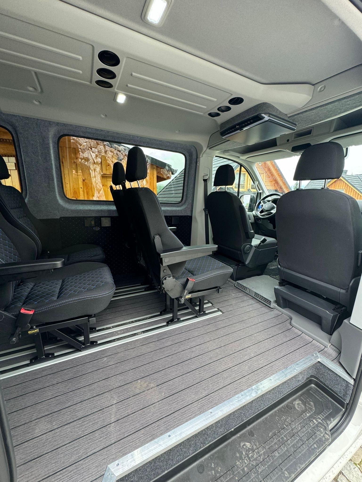 Przestronne wnętrze busa z regulowanymi fotelami na szynach, wykończone szarą wykładziną imitującą drewno i jasną tapicerką. Widok z tyłu kabiny pasażerskiej.