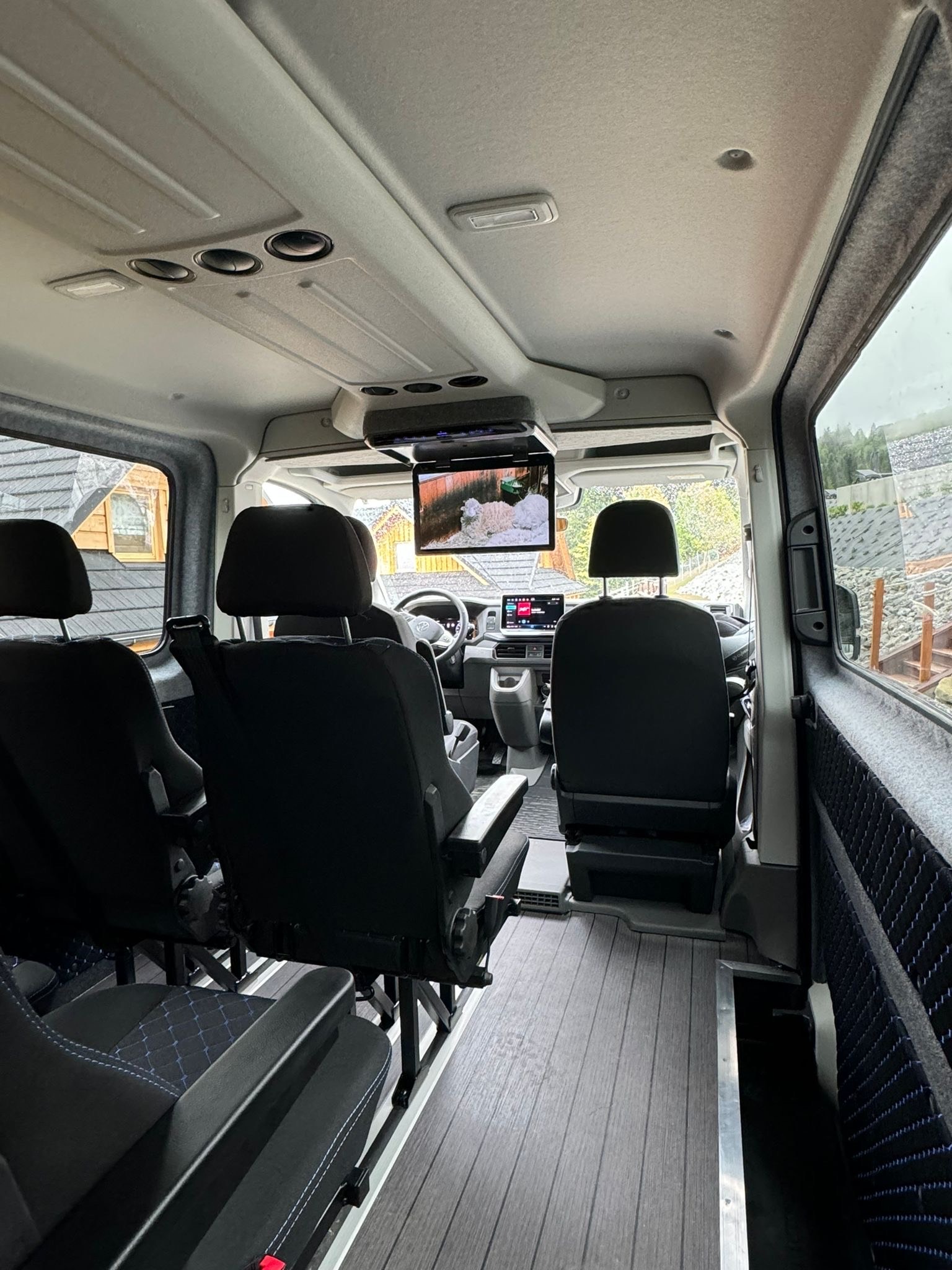 Wnętrze luksusowego busa z czarnymi skórzanymi siedzeniami, drewnianą podłogą i ekranem TV zamontowanym pod sufitem, widok z tyłu pojazdu.