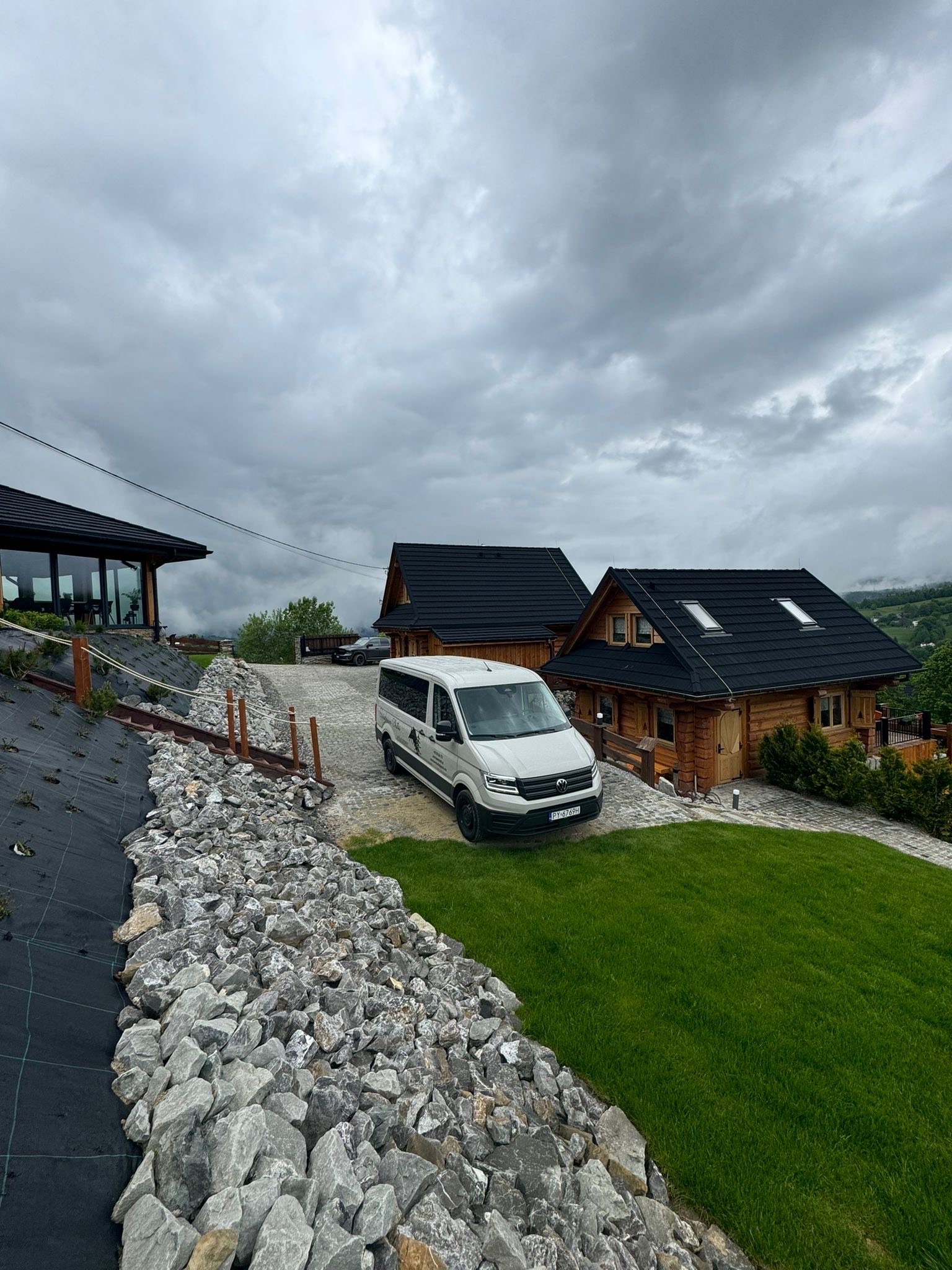 Jasny bus na tle drewnianych domków z czarnymi dachami, otoczonych zielenią i kamieniami. Pochmurne niebo. Widok z dołu, budzący zaufanie.