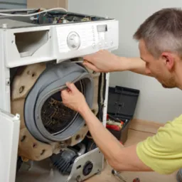 Serwisant w Rudzie Śląskiej naprawia pralkę, odkręcając śrubokrętem gumowy kołnierz bębna. Widoczna skrzynka z narzędziami i otwarte drzwiczki pralki.