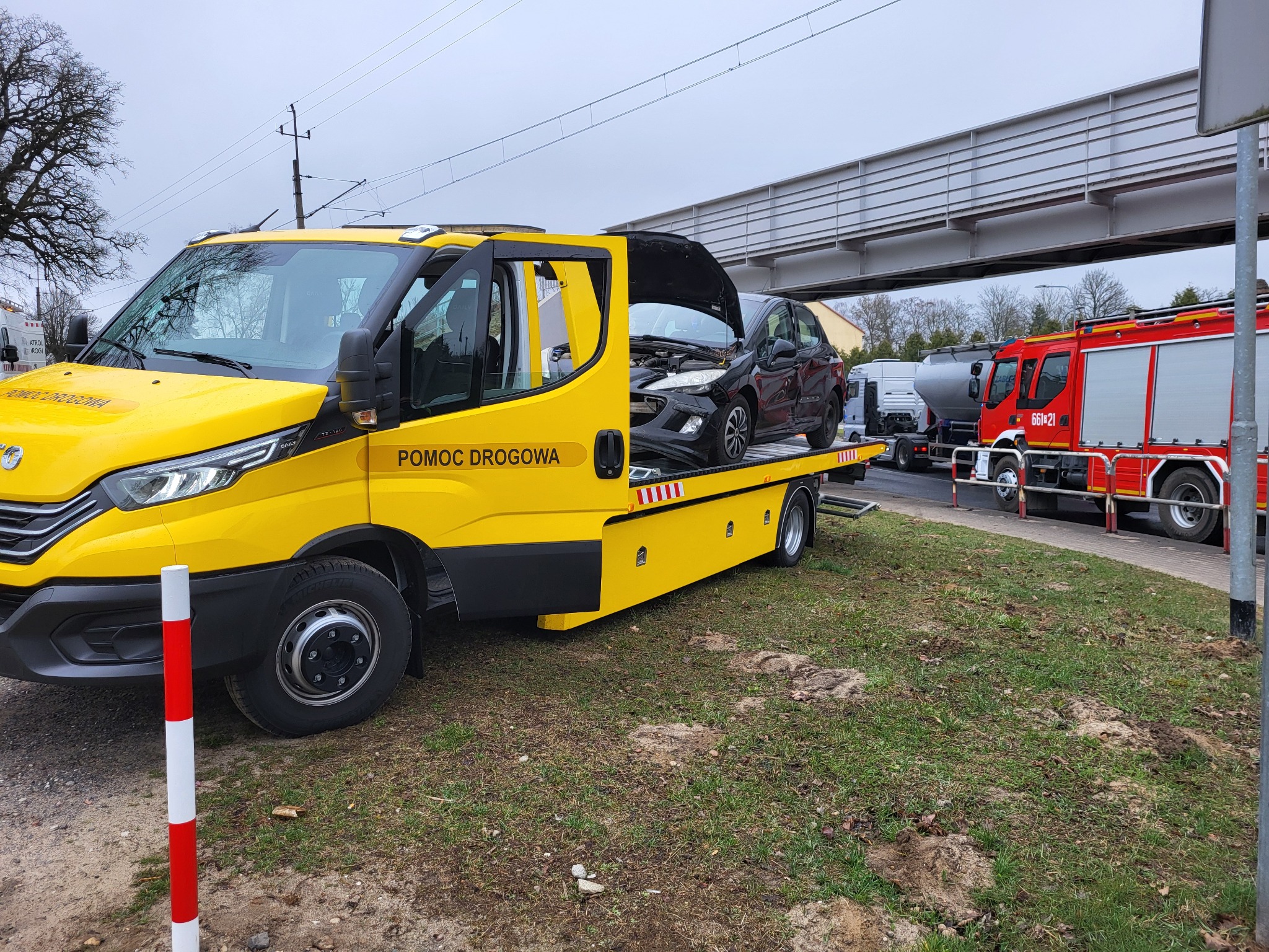 Żółta laweta z uszkodzonym czarnym autem na platformie, w tle wóz strażacki i wiadukt. Widoczny napis 'Pomoc Drogowa'.