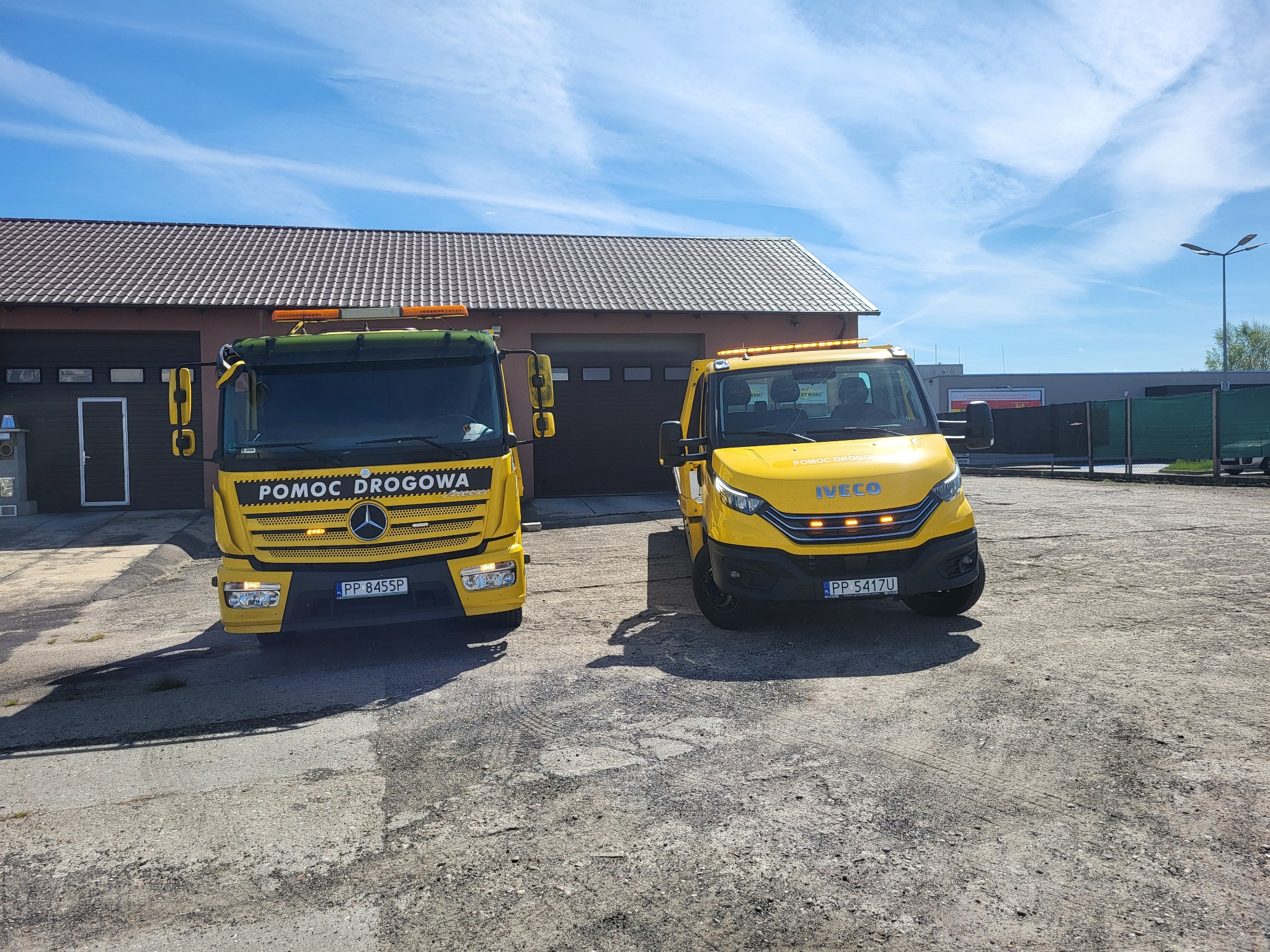 Dwa żółte pojazdy pomocy drogowej, Mercedes i Iveco, stojące na betonowym placu przed budynkiem z brązowym dachem w słoneczny dzień.