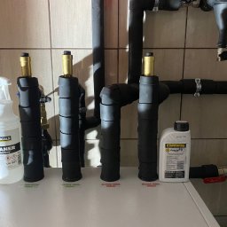 TUTTO SPÓŁKA Z OGRANICZONĄ ODPOWIEDZIALNOŚCIĄ - Fragment instalacji grzewczej z rurami w izolacji, z widocznymi etykietami 'wejście' i 'wyjście'. Obok stoją preparaty do czyszczenia i ochrony instalacji.