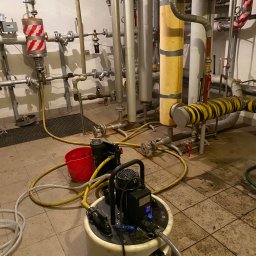 TUTTO SPÓŁKA Z OGRANICZONĄ ODPOWIEDZIALNOŚCIĄ - Pompa zanurzeniowa w kotłowni, podłączona do węży. W tle rury instalacji grzewczej z izolacją w kolorach czerwono-białym i żółto-czarnym.