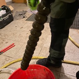 TUTTO SPÓŁKA Z OGRANICZONĄ ODPOWIEDZIALNOŚCIĄ - Czyszczenie rury spiralną sprężyną hydrauliczną nad czerwonym wiadrem z brudną wodą. W tle narzędzia i elementy instalacji.