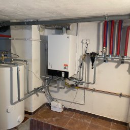 TUTTO SPÓŁKA Z OGRANICZONĄ ODPOWIEDZIALNOŚCIĄ - Biały piec gazowy Viessmann z szarymi rurami, czerwono-niebieskimi izolacjami i zbiornikiem na wodę w kotłowni, na tle beżowej ściany i brązowej podłogi.