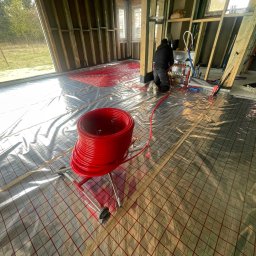 TUTTO SPÓŁKA Z OGRANICZONĄ ODPOWIEDZIALNOŚCIĄ - Instalacja ogrzewania podłogowego w surowym wnętrzu. Widoczne czerwone rury na folii z siatką, rozdzielacz i instalator w trakcie pracy. Surowy stan budynku.