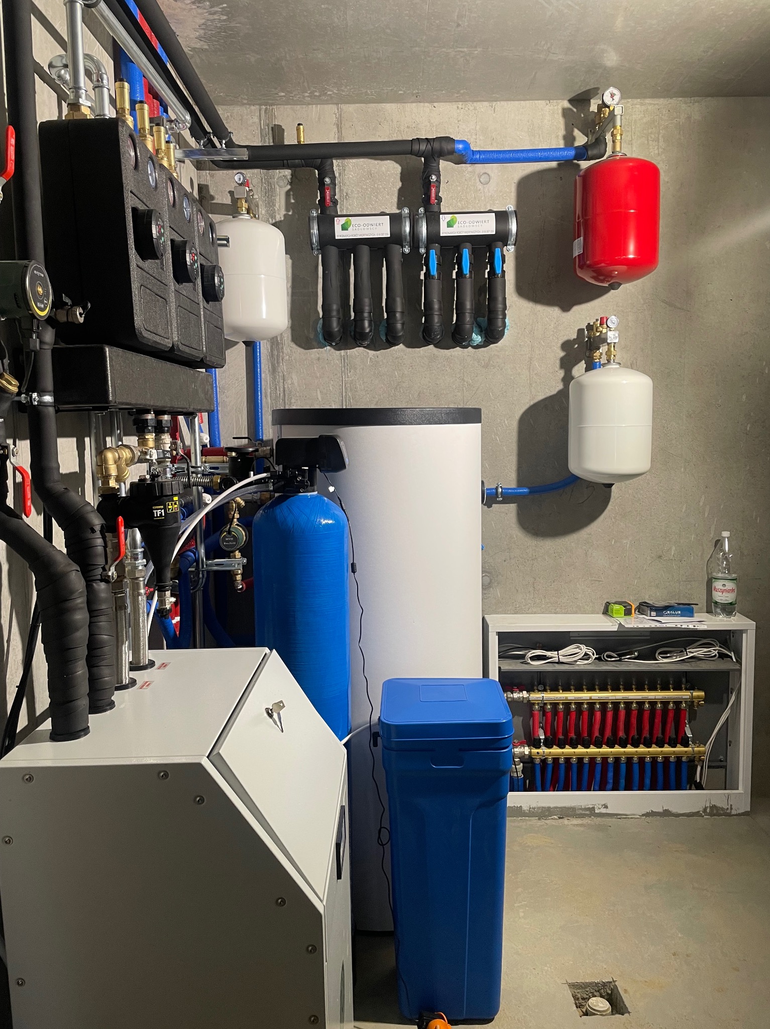 Kompletna kotłownia z filtrami, zbiornikami, rozdzielaczem ogrzewania podłogowego i kotłem. Widok na całą instalację w pomieszczeniu technicznym.