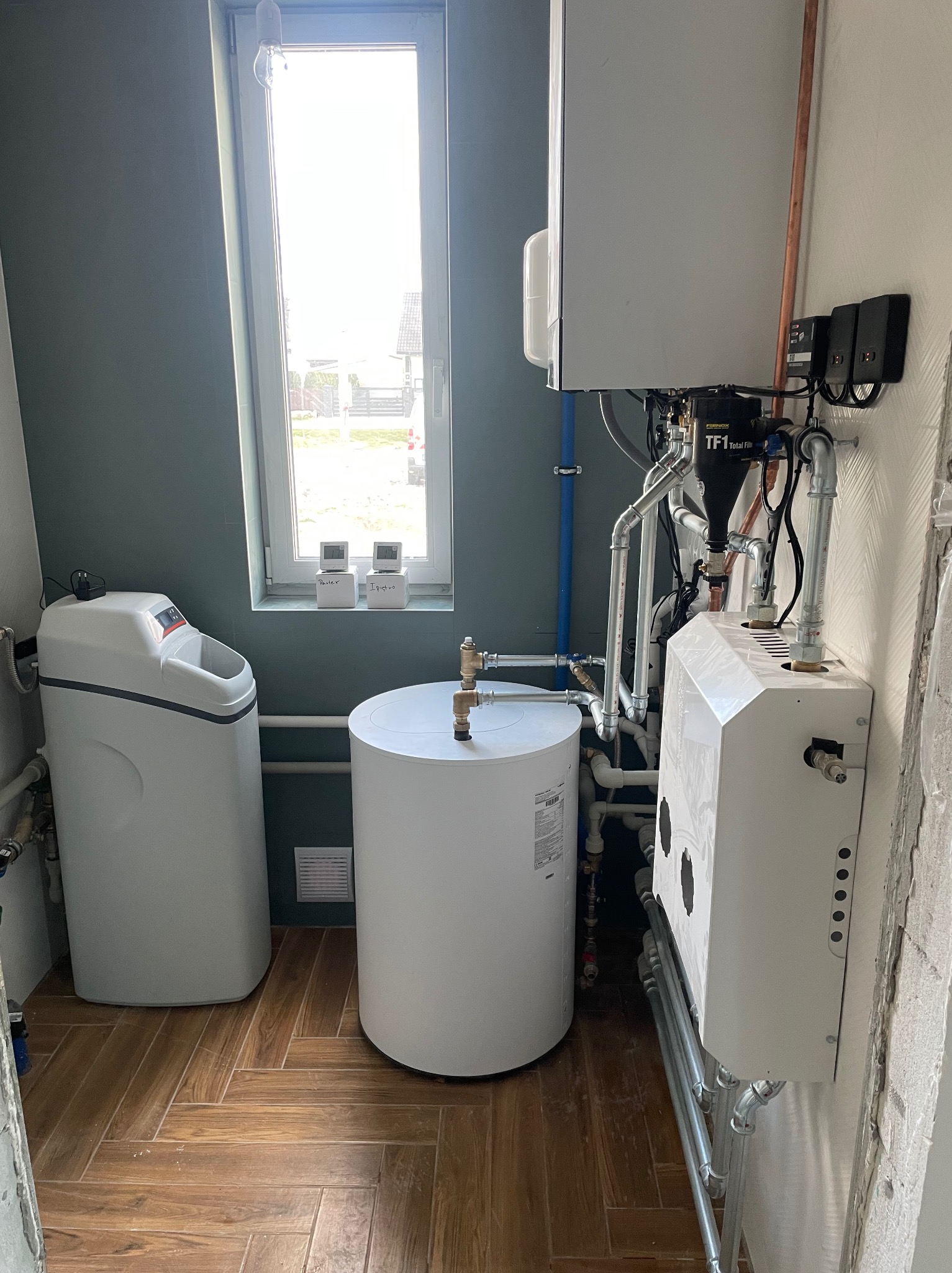 Kompletna instalacja CO w kotłowni: piec gazowy, zbiornik buforowy, zmiękczacz wody, rury miedziane. Widok na okno z parapetem z dekoracjami.