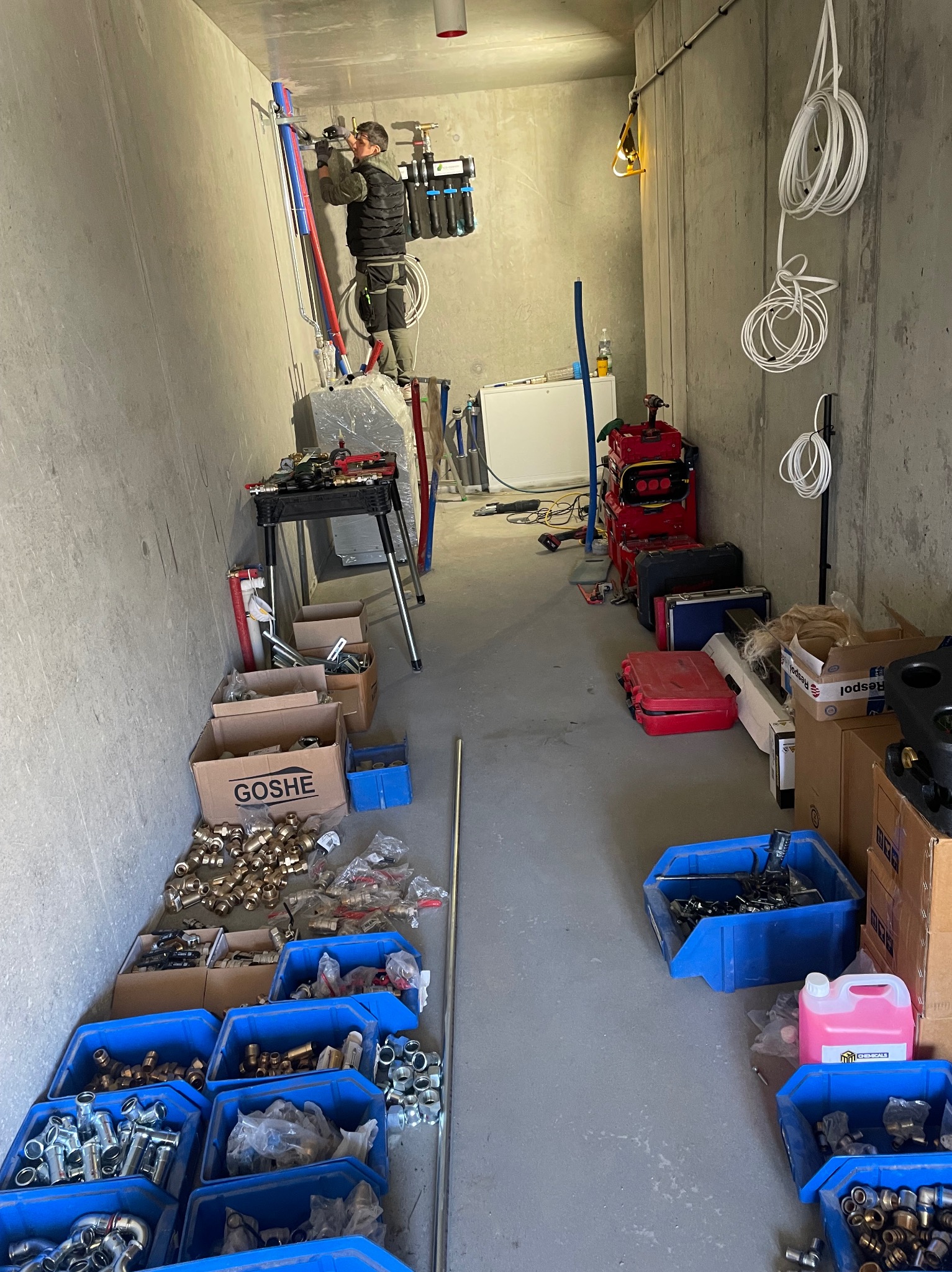 Instalator na drabinie montuje instalację hydrauliczną w surowym wnętrzu. Widoczne liczne akcesoria, rury i narzędzia. Oświetlenie robocze.